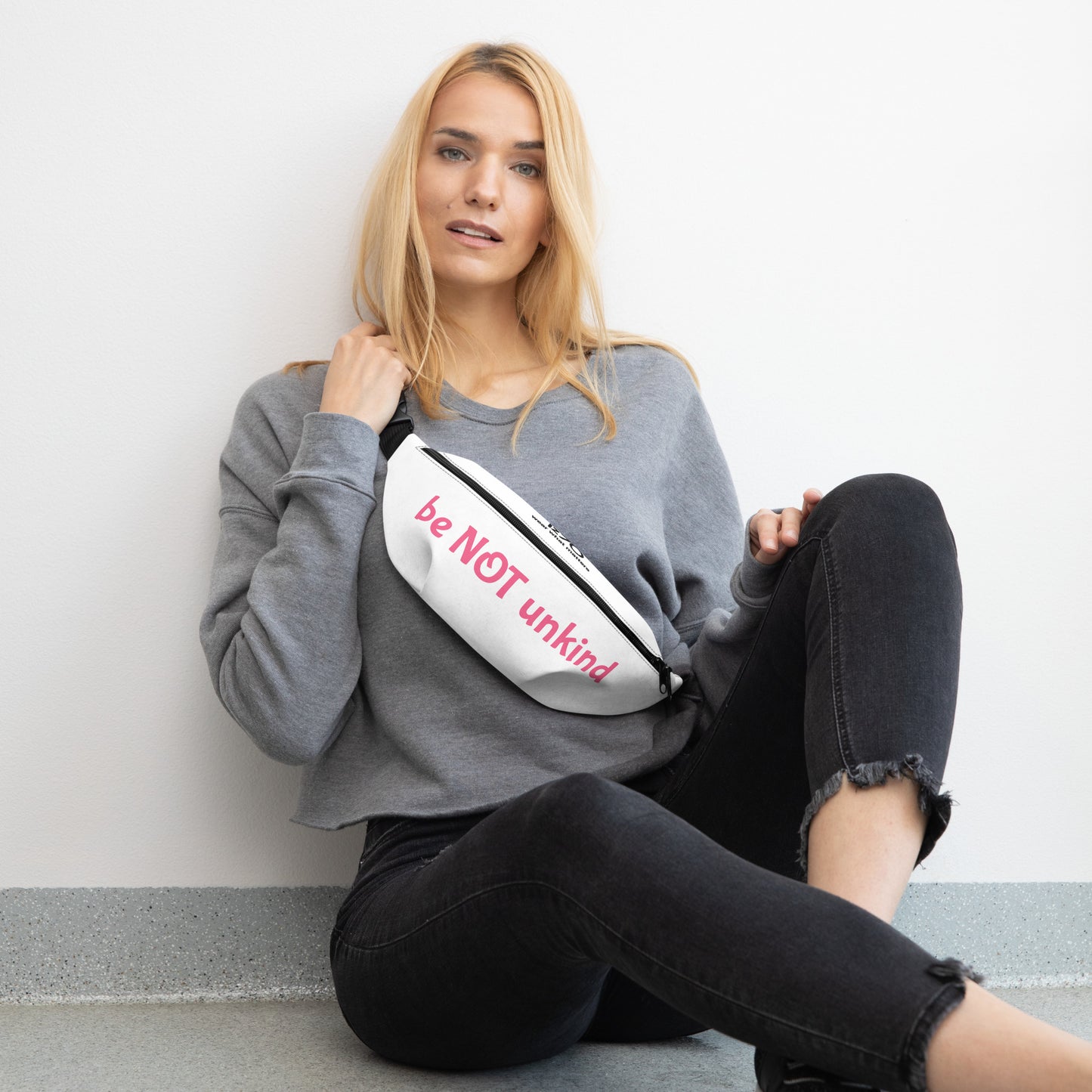 Woman sitting on the floor holding a white fanny pack with text, wearing a gray sweatshirt and black pants.