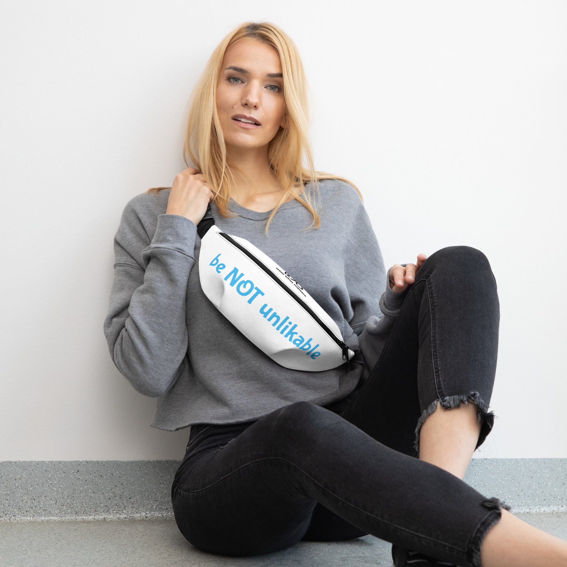 Woman sitting on the floor wearing a gray sweatshirt and black pants, holding a white fanny pack with text.