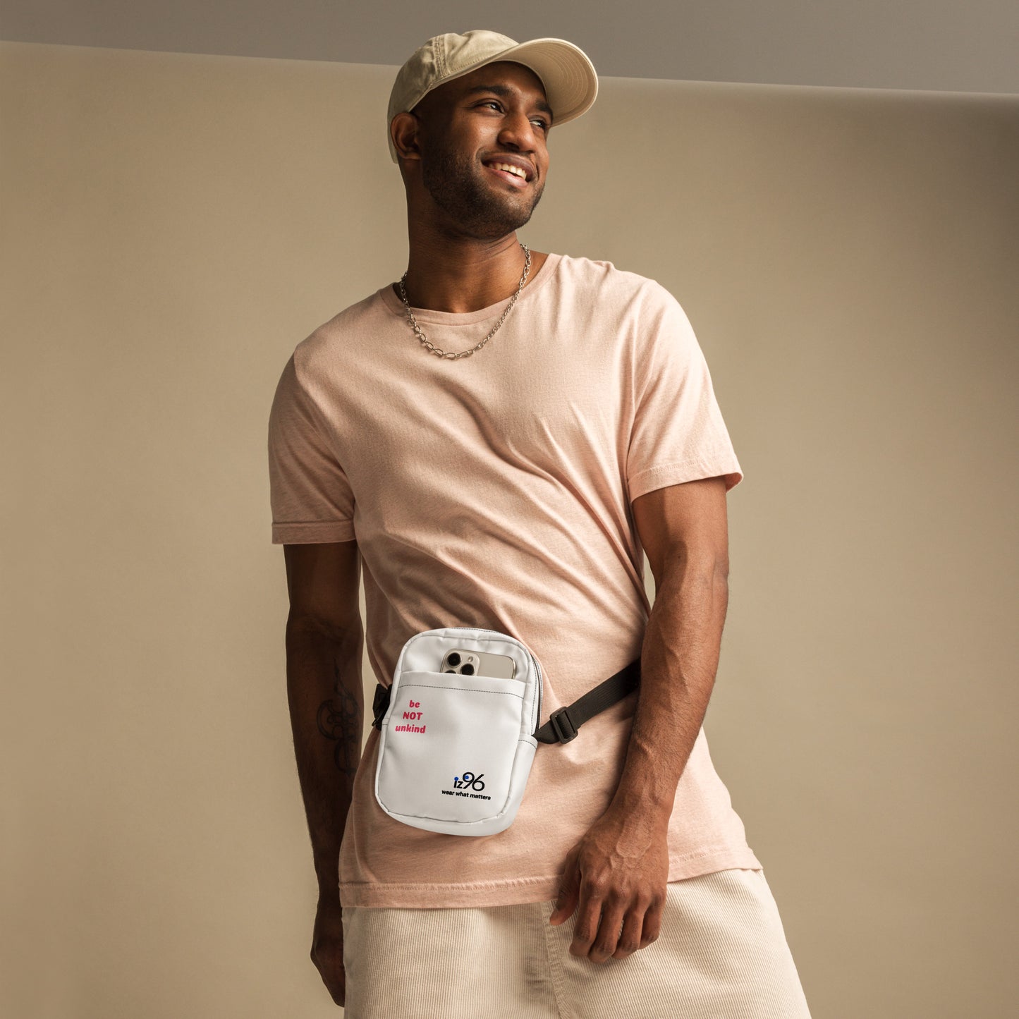 Man wearing a light pink t-shirt and beige cap, holding a white waist bag with a logo against a beige background