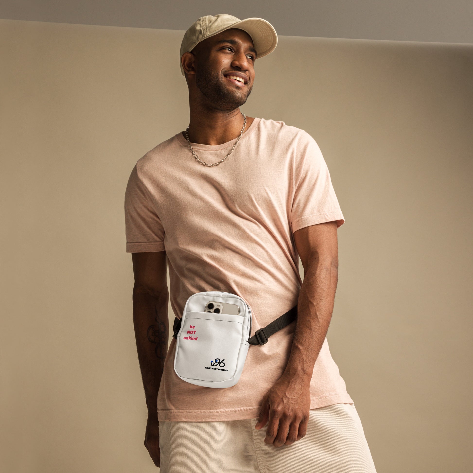 Man wearing a light pink t-shirt and beige cap, holding a white waist bag with a logo against a beige background