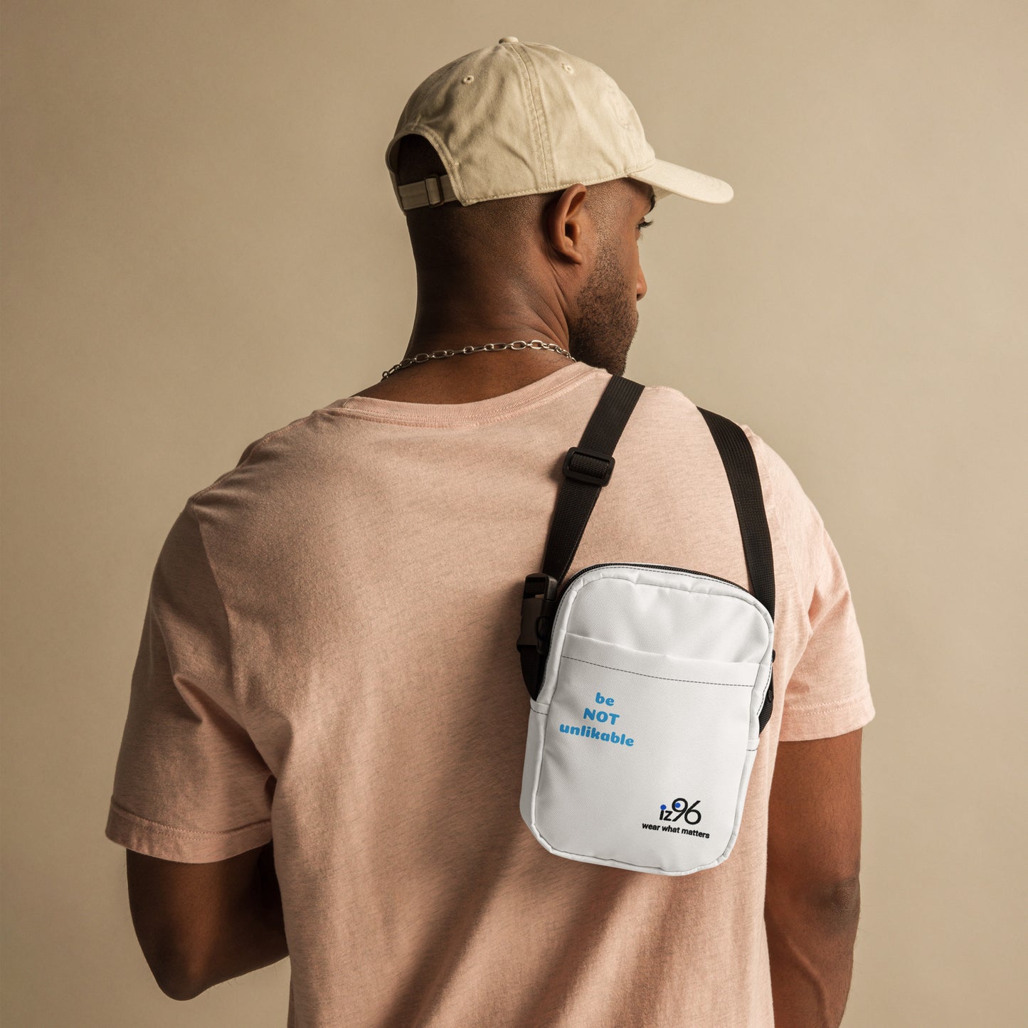 Man wearing a beige cap and light pink shirt with a white crossbody bag on a beige background
