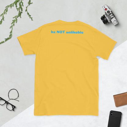 Yellow t-shirt with 'be NOT unlikable' text on a light gray surface with accessories.