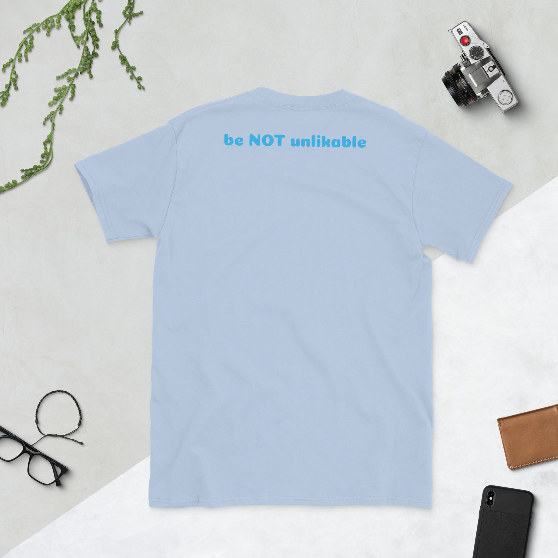 Light blue t-shirt with 'be NOT unlikable' text on a light gray surface with accessories.