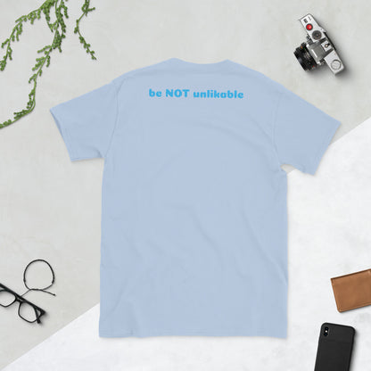 Light blue t-shirt with 'be NOT unlikable' text on a light gray surface with accessories.
