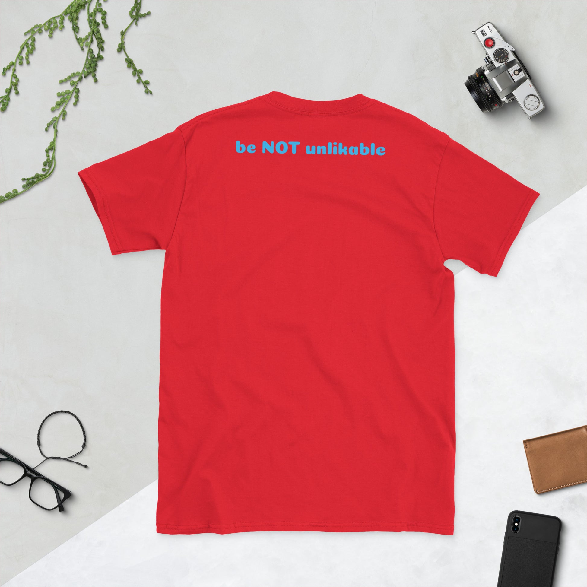 Red t-shirt with 'be NOT unlikeable' text on a light gray surface with accessories.