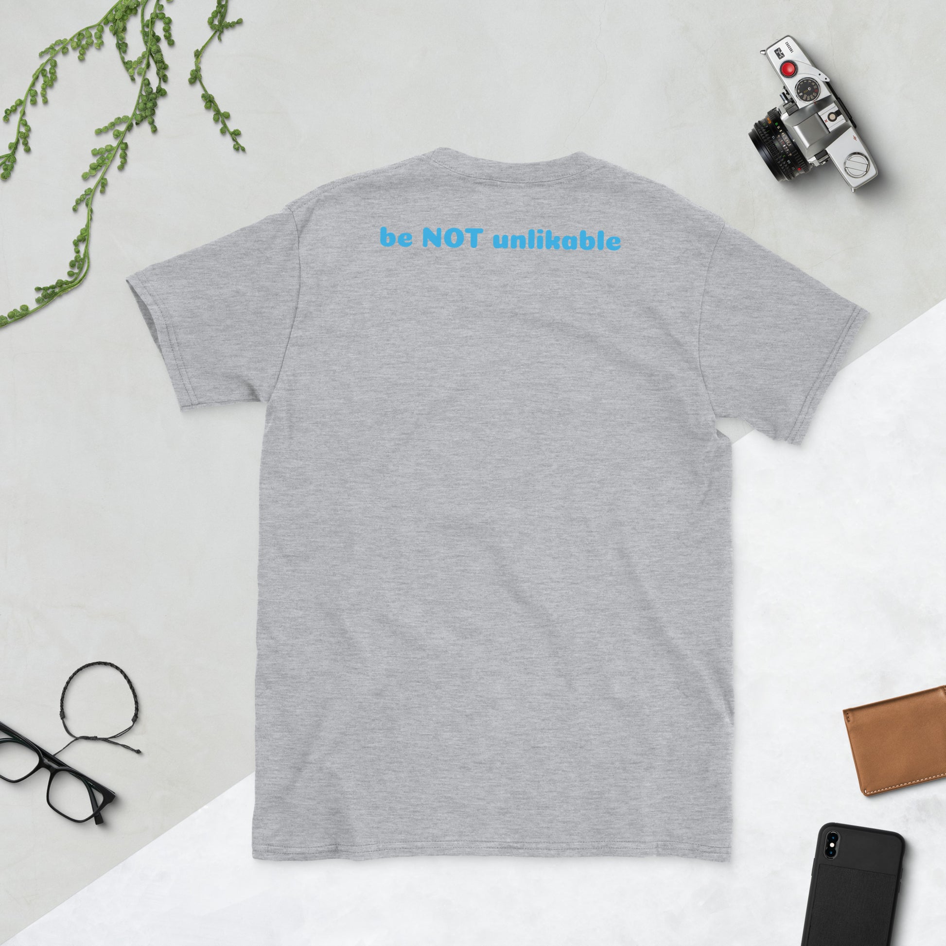Gray t-shirt with 'be NOT unlikable' text on a light gray background with accessories.