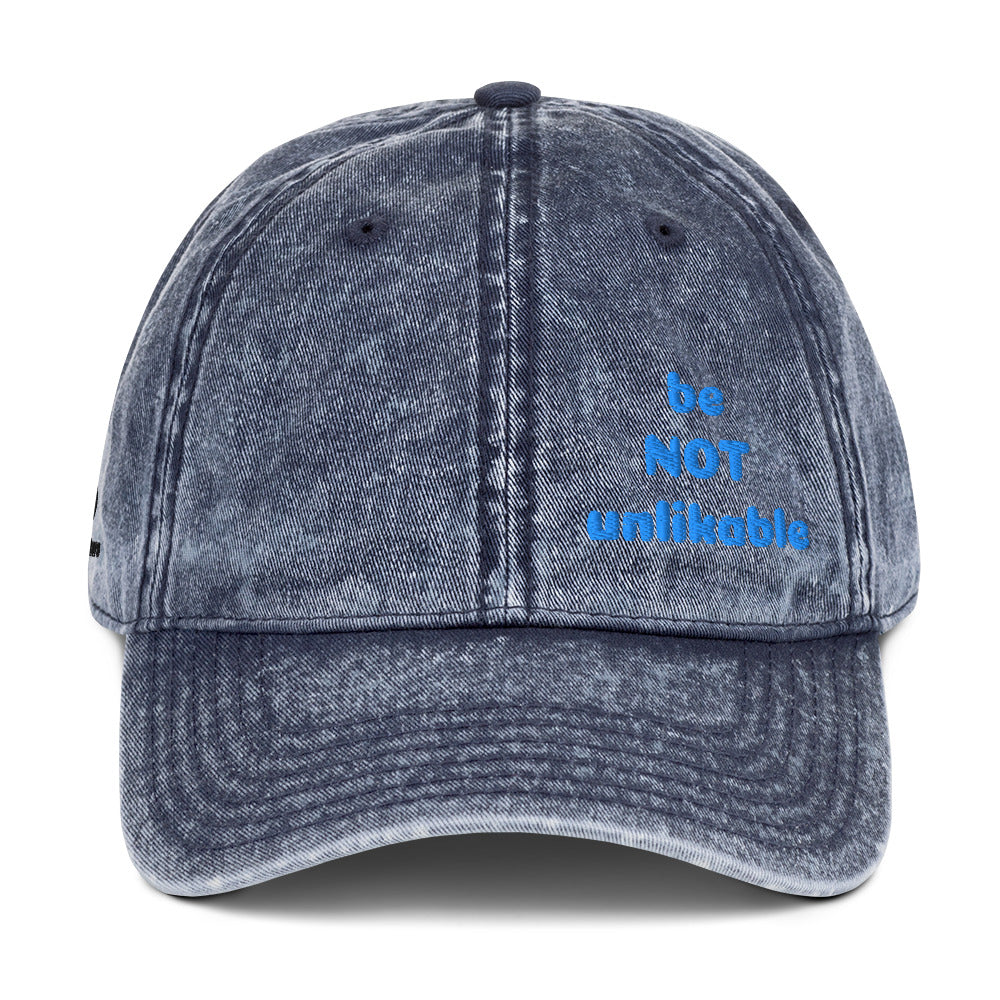 Blue denim cap with 'be NOT unlikable' text on a white background