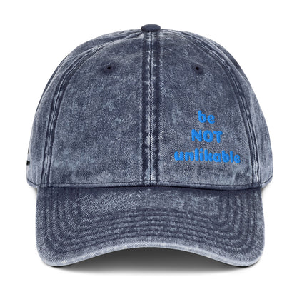 Blue denim cap with 'be NOT unlikable' text on a white background