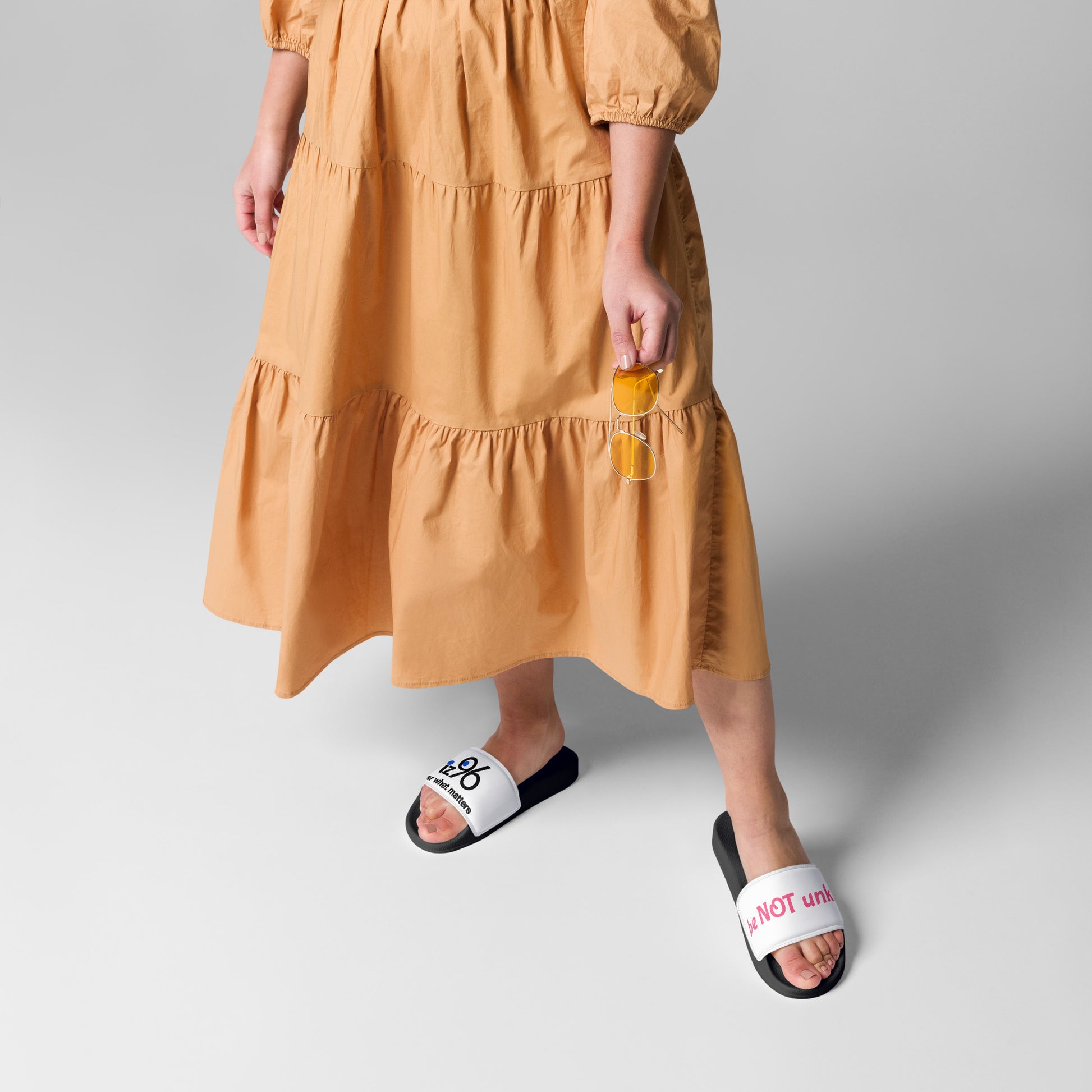Person wearing a mustard yellow dress and black slides with white soles on a gray background
